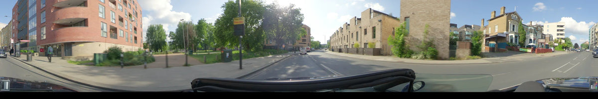 Past modern brick housing and green trees, a car follows a white van on a partly cloudy spring day in London, United Kingdom.