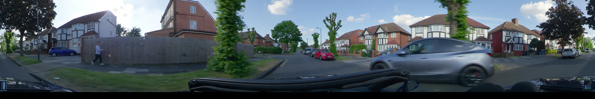 Passing brick houses and lush green trees, a car drives through a residential street in London, United Kingdom, on a clear spring day.