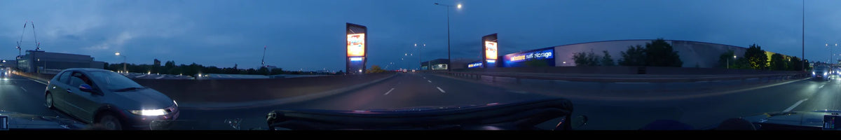 Bright digital billboards illuminate a road in London, United Kingdom, while a car drives past a storage facility on an overcast spring evening.