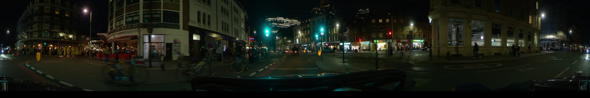 Passing green traffic lights and lit storefronts, a car travels through London, United Kingdom, during a clear spring night with a brightly illuminated building ahead.