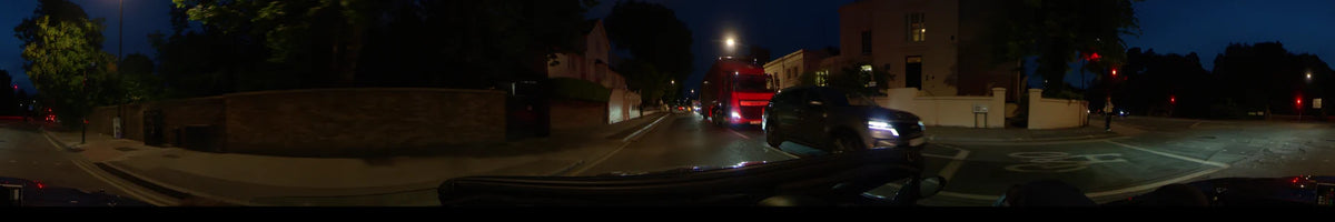 Under glowing street lamps, a car follows a large red truck through a residential neighborhood in London, United Kingdom, during an overcast spring night.