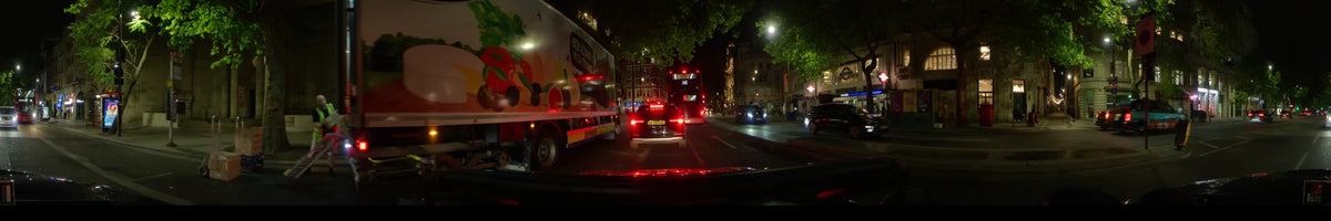 A red double-decker bus and a car lead the way past a fruit-themed truck along a road in London, United Kingdom, during a clear spring night.