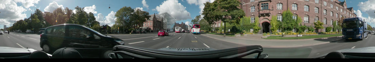 A wide city street in Copenhagen featuring a mix of historic brick houses and a modern geometric building, with buses and a red car in traffic.