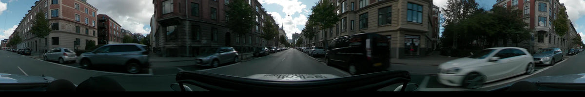 Historic brick apartment buildings and green trees line a city street in Copenhagen, with parked cars and a tall office building visible in the distance.