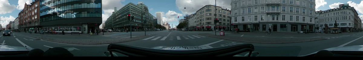 A wide city road in Copenhagen with a pedestrian crossing, flanked by multi-story residential buildings and a tall white office tower under a partly cloudy sky.