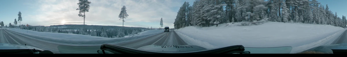 A car drives through a snowy winter landscape in Dalarna, Sweden, during a partly cloudy sunrise. Snow-covered pine trees line the road as another vehicle approaches.