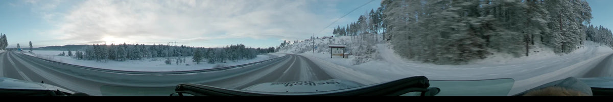 A car drives through a snowy landscape in Idre, Dalarna, Sweden, during a partly cloudy sunrise in winter, passing a road sign and snow-covered hills.