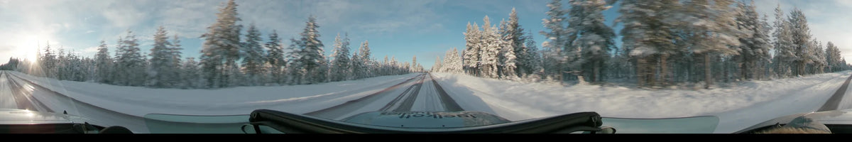 Snow-laden pine trees flank a winter road in Dalarna, Sweden, as a car drives under a partly cloudy sky during the day.
