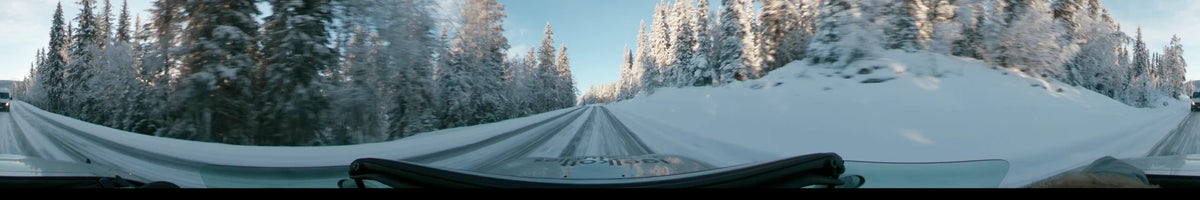 A car traverses a snowy road in Dalarna, Sweden, passing through dense, snow-covered pine forests during a partly cloudy winter day.