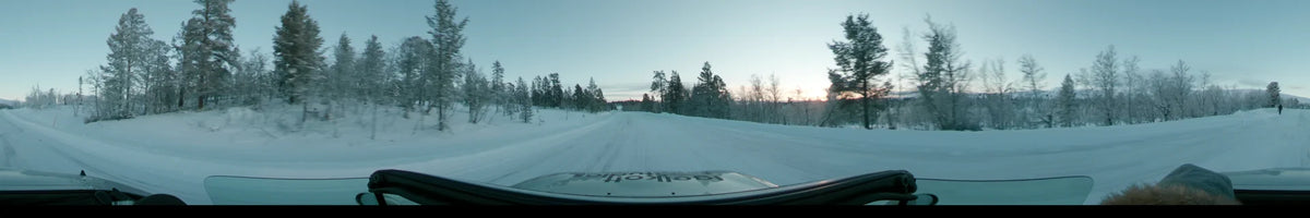 In Grövelsjön, Sweden, a car travels a snowy road lined with frosted trees under a partly cloudy sky during a winter dusk.