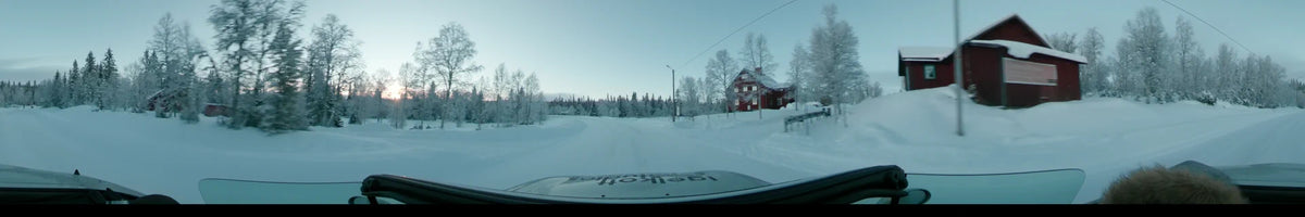 Passing a red house and frosted trees, a car travels along a snowy road in Grövelsjön, Sweden, during a partly cloudy winter dusk.