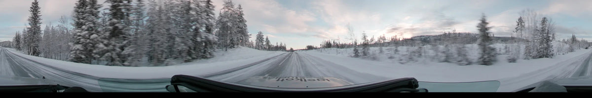 A car drives along a snowy road in Grövelsjön, Sweden, during a partly cloudy winter afternoon, surrounded by snow-covered pine forests.