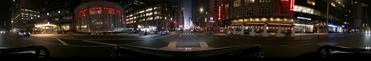 A car drives through New York City at night during spring. The scene features tall buildings, bright neon signage, and city traffic along a wide urban road.