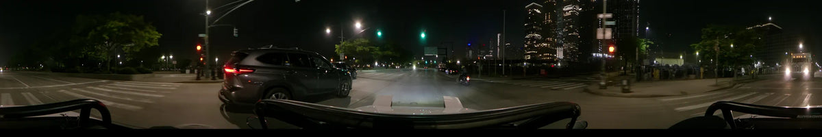 Green traffic signals and city lights illuminate a New York street in the United States as a car drives past other vehicles and a motorcycle on a spring night.