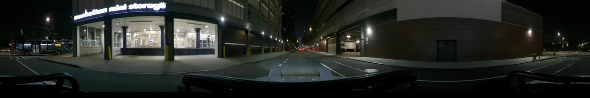 Industrial warehouses with loading docks and yellow bollards line a New York road at night during spring, seen from a car's point of view.