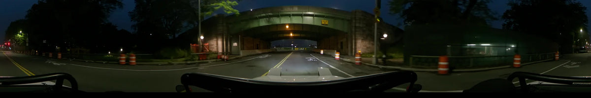 A car travels through a New York underpass past orange construction barrels beneath a green metal ceiling on a spring night in the United States.