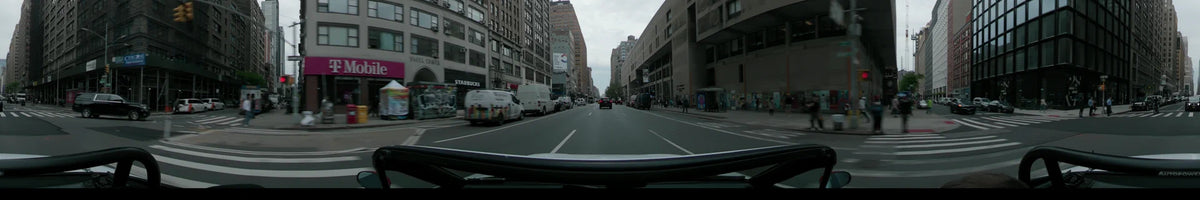 Daytime car POV driving through New York City during spring. The scene features an overcast sky, tall urban buildings, and several white delivery vans along the road.