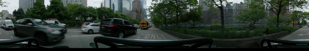A yellow school bus and black SUV lead a car through New York on an overcast spring day, with One World Trade Center towering in the background.