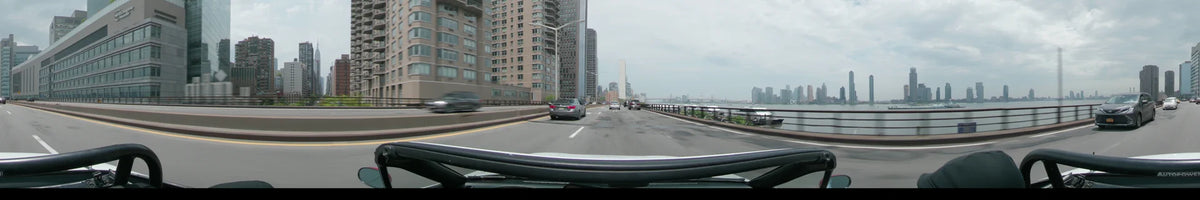 High-rise apartments and a river view accompany a car driving through New York on a partly cloudy spring day, with distant bridges crossing the water.
