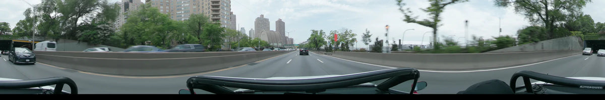 On a partly cloudy spring day in New York, a black SUV travels ahead of the car past a large arched structure and towering brick high-rises.