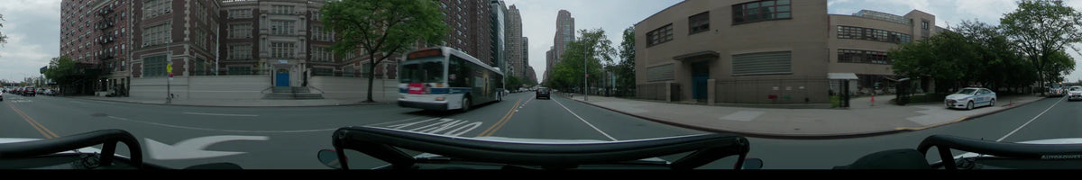 Passing a white transit bus on a partly cloudy spring day, a car follows another vehicle along a New York road lined with tall brick apartments and trees.