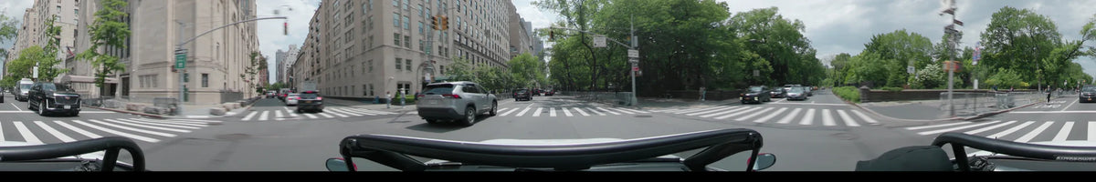 Tall apartment buildings and lush green trees flank a New York road where a car follows a silver SUV on a partly cloudy spring day.