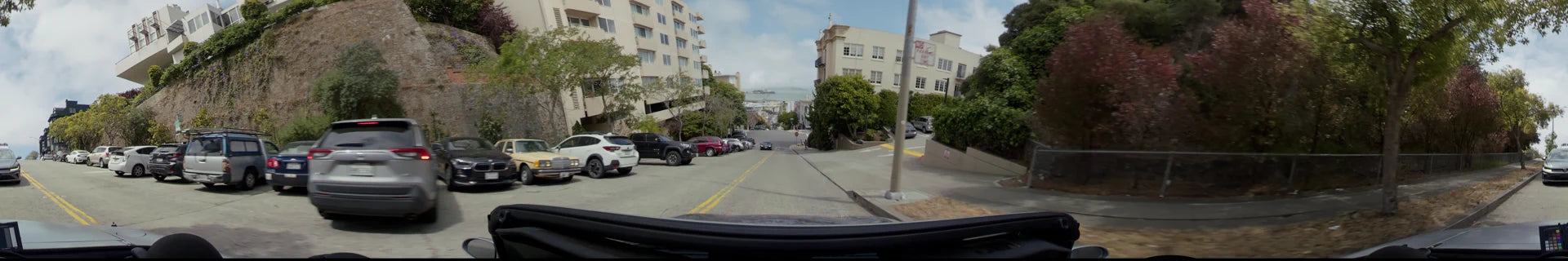 A residential street in San Francisco with parked cars and multi-story apartment buildings, leading toward a distant view of the bay.