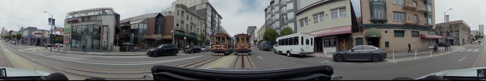 Two iconic cable cars travel along a track-lined city street in San Francisco, flanked by multi-story residential buildings, parked cars, and a passing white bus.