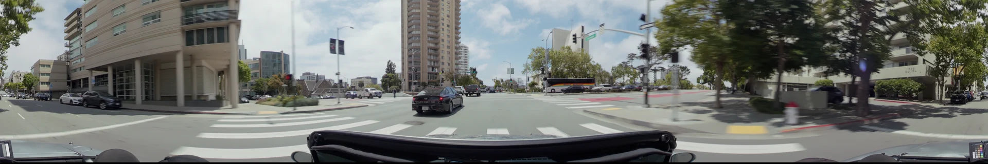 A wide city intersection in San Francisco with a black sedan and a large tour bus passing high-rise residential buildings and green city trees.