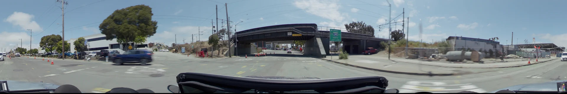 A road in San Francisco passing under a dark industrial bridge with height clearance signs, orange traffic cones, and a white car driving ahead.