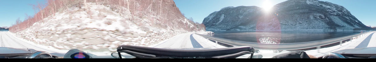 View from a car driving along a snow-covered road in Stavanger, Norway. On a clear spring day, the path curves beside a fjord and snowy mountains.