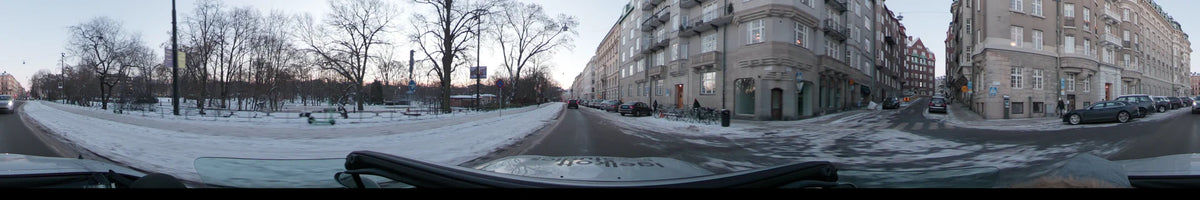 A city street in Stockholm, Sweden, features apartment buildings and snow-covered sidewalks as a car drives past bare trees during a clear winter dawn.
