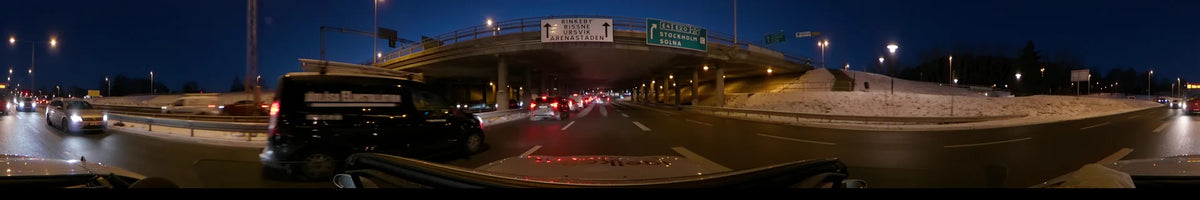 Concrete underpasses and road signs for Solna and Rinkeby surround a car driving through city traffic in Stockholm, Sweden, during a clear winter sunset.