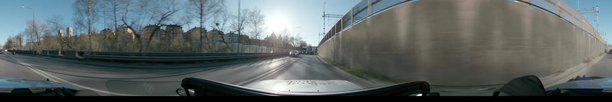 Sunlight glares as a car follows traffic past a concrete sound wall and distant city buildings on a clear spring day in Stockholm, Sweden.