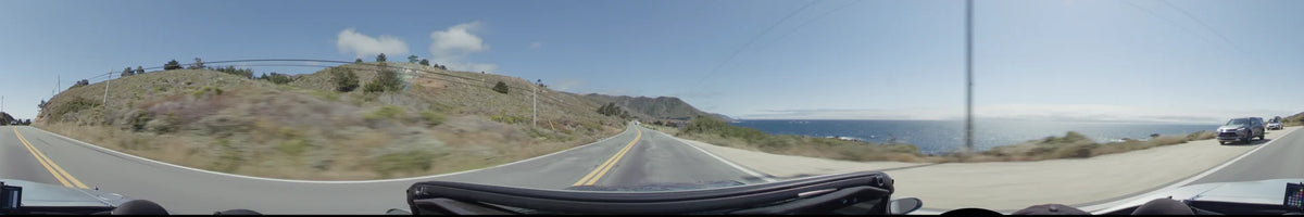 A winding two-lane highway in Big Sur, flanked by rugged coastal hills and the blue ocean under a clear sky.