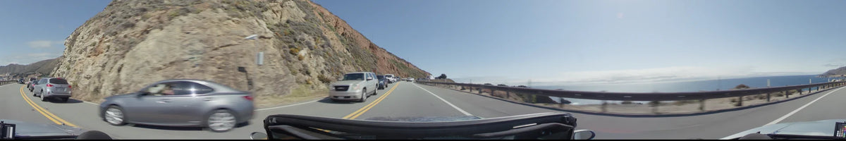 A line of cars travels along a coastal highway in Big Sur, flanked by steep, rocky cliffs and a roadside guardrail under a clear sky.