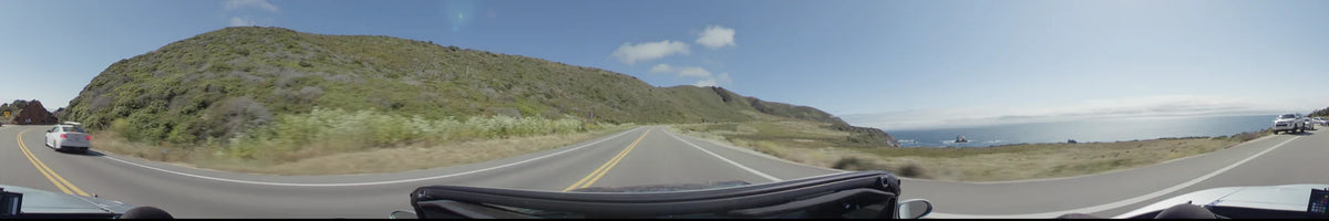 A two-lane road curves through Big Sur, bordered by steep, green-covered slopes and rolling hills under a bright, clear sky.