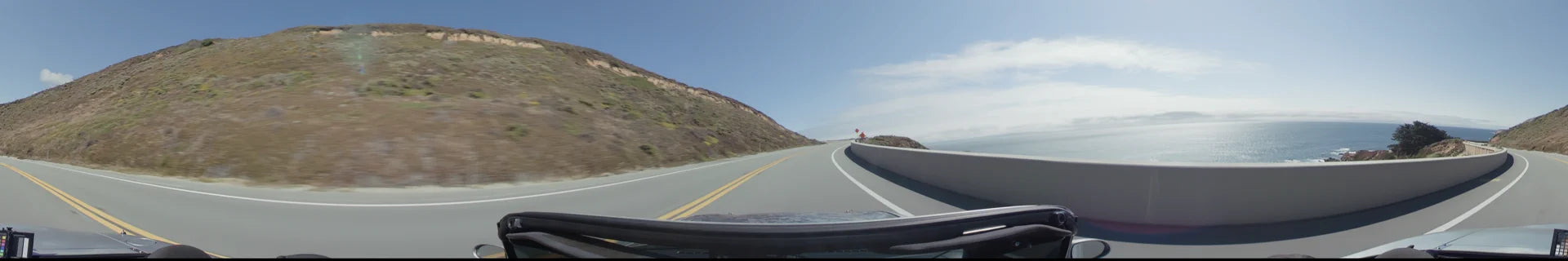 A curving coastal road in Big Sur follows a steep, rocky hillside, bordered by a concrete barrier and bright yellow center lines under a clear sky.