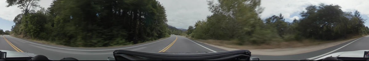 A two-lane road curves through a dense forest of tall green trees in Big Sur, with bright yellow center lines leading toward distant mountains.