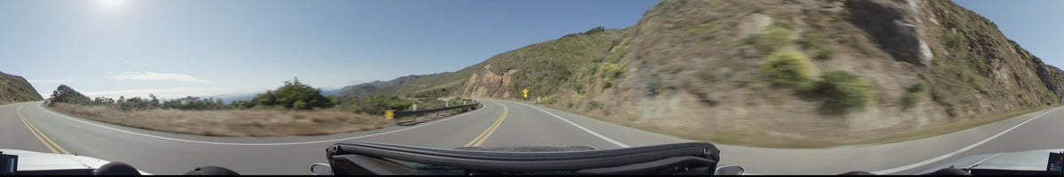A curving road in Big Sur winds past steep, rocky cliffs and brush-covered hills, marked by yellow center lines and a roadside warning sign.