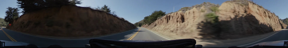 A two-lane road in Big Sur cuts through steep, sandy cliffs and scrubland, with yellow center lines leading toward distant green hills under a clear sky.