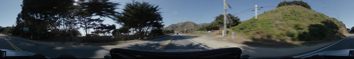 A straight road in Big Sur leads toward rolling green mountains, flanked by tall evergreen trees and a roadside fence under a clear afternoon sky.