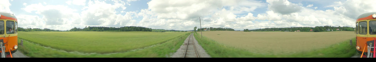 View from a train passing through green summer fields in Uppsala, Sweden, under a partly cloudy sky with utility poles and distant houses along the tracks.