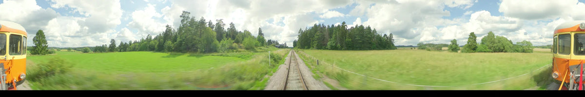 View from a train traveling along straight tracks in Uppsala, Sweden. Green summer meadows and forests line the route under a partly cloudy day sky.