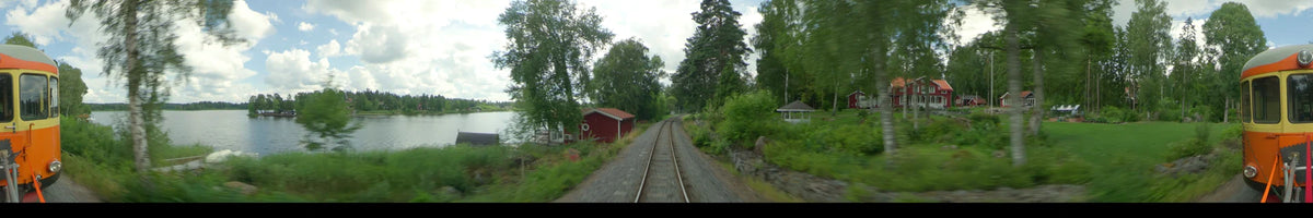 View from a train moving through a lush summer landscape in Uppsala, Sweden. A red cottage and a lake are visible under a partly cloudy sky.