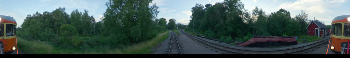 View from a train in Uppsala, Sweden, during a partly cloudy summer afternoon. The railway tracks diverge through lush green foliage with a small building in the distance.