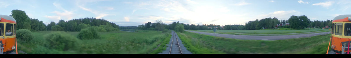 View from a train moving through the summer landscape of Uppsala, Sweden, on a partly cloudy afternoon, passing by green fields, trees, and a road.