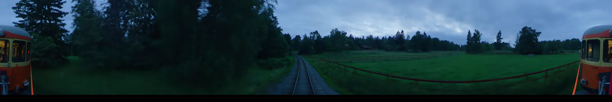View from a train traveling through the summer landscape of Uppsala, Sweden. The tracks curve past green forests and a red fence under a partly cloudy afternoon sky.