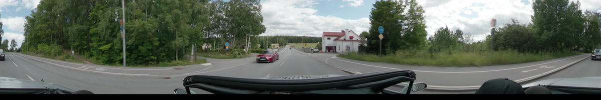 A red car drives along a road in Uppsala, passing a white building with a red roof and lush green trees under a partly cloudy sky.