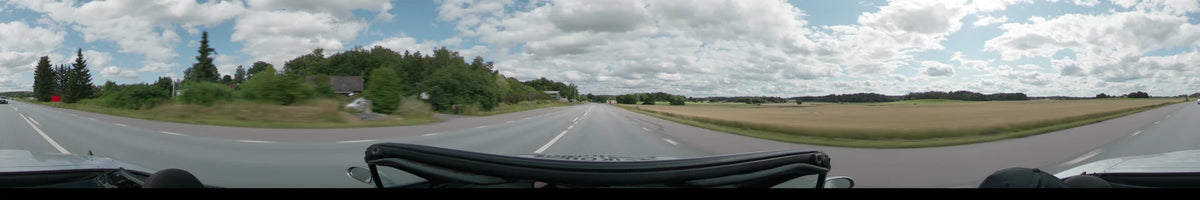 A straight road between Uppsala and Upplands-Bro is flanked by golden fields and lush green trees under a partly cloudy summer sky.